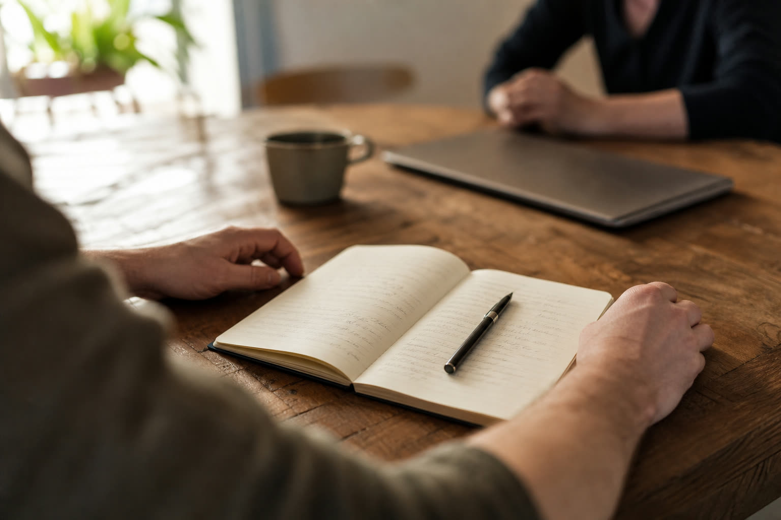 An open notebook of handwritten notes on a meeting table, pen resting on the page, a second person out of focus across the table.